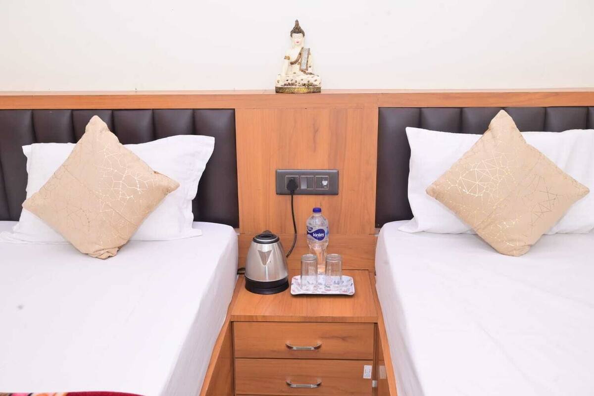 Habitación de hotel con dos camas, cada una con una almohada blanca y una almohada color beige, una pequeña estatua en la cabecera, una botella de agua y una tetera en la mesita de noche.
