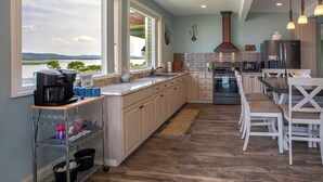 A modern kitchen with a view of a lake, a coffee maker, and a dining area with white chairs. - Riverview, 5BR, Hot tub, Fireplace, Arcade Game (Minneiska)