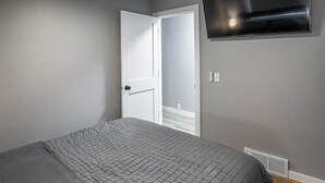 A bedroom with a bed, a white door, a wall-mounted TV, and a vent.