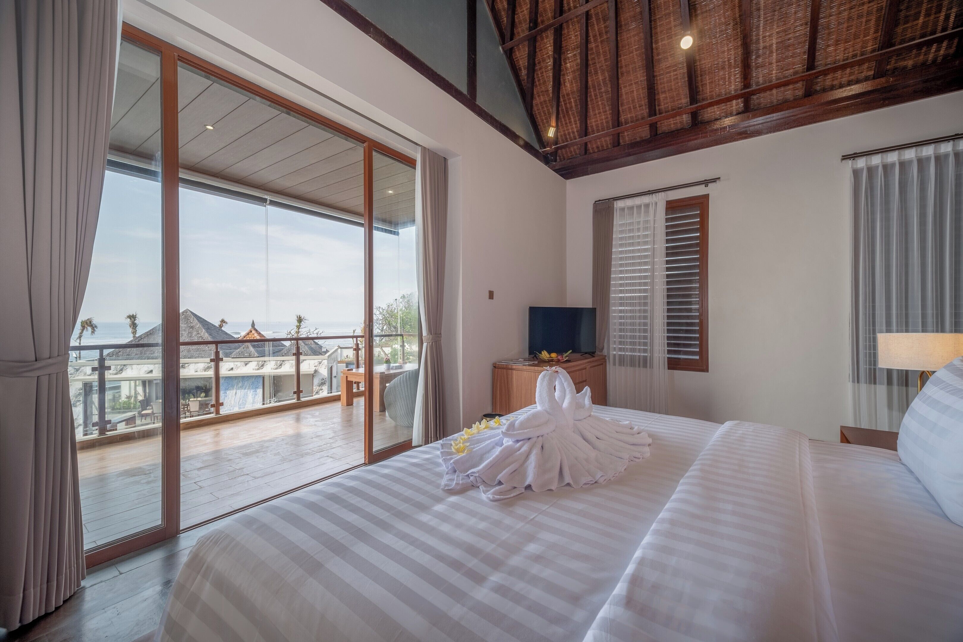 A hotel room with a large bed, a TV, a wooden nightstand, and a balcony with a view of the resort.