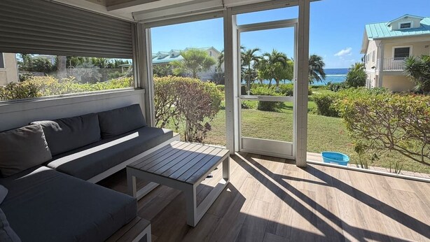 A modern outdoor seating area with a sofa and table, large windows, and a view of the ocean and greenery. - Cayman Beach Escape - luxury ground floor 2BR (Savannah)
