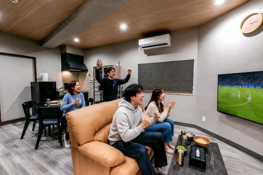 A modern living room with a large flat-screen TV, a sofa, a coffee table with snacks, and a dining area with chairs and a table.