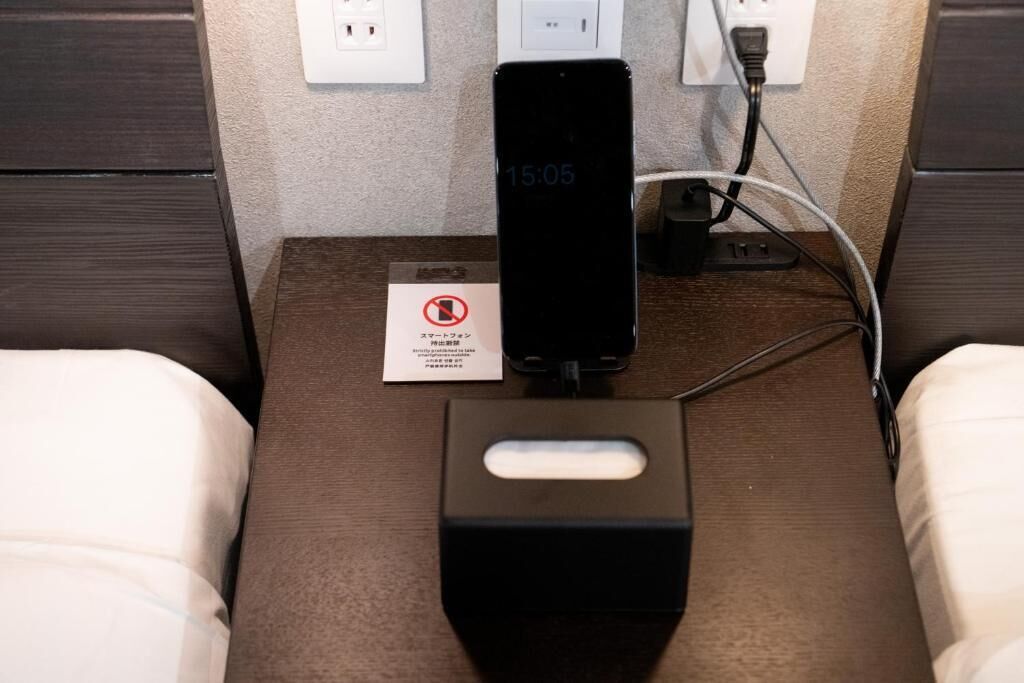 A smartphone charging on a bedside table with a power strip, a tissue box, and a no-smoking sign.