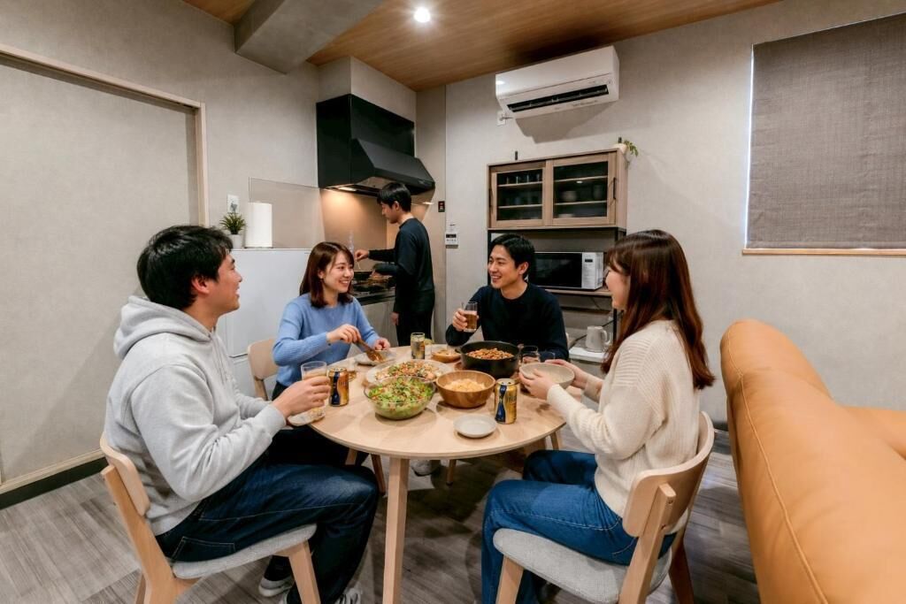 A group of people dining around a round table with various dishes and drinks, in a room with a modern kitchen and a large window.