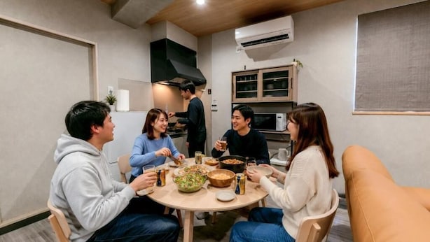 Un grupo de personas cenando alrededor de una mesa redonda, con varios platos y bebidas, en una habitación con una cocina moderna y un ventanal grande.
