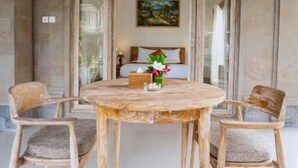 A room with a round wooden table, four chairs, a painting on the wall, and a bed in the background. - Lebah Sari House (Ubud)