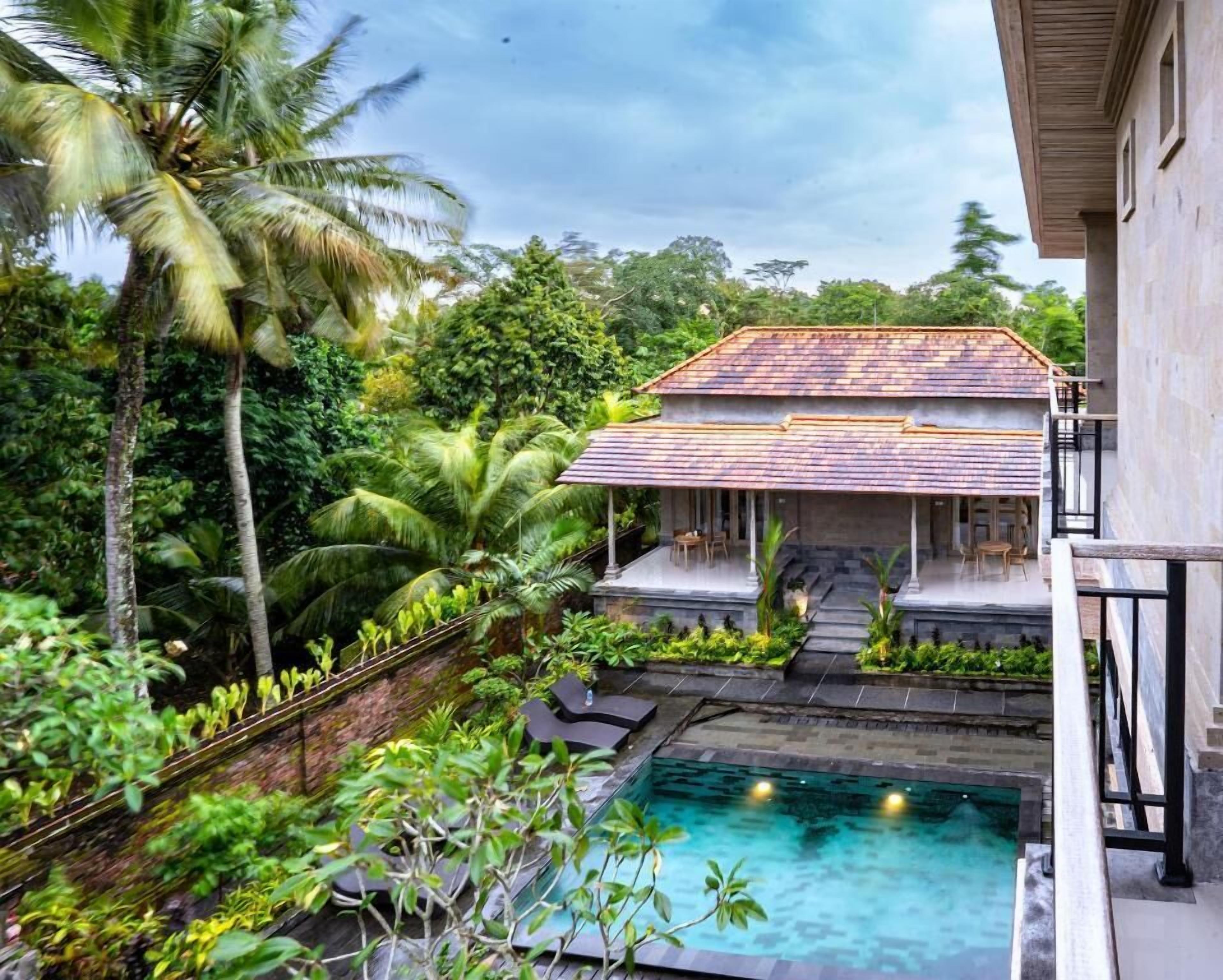 A pool with a stone edge, a house with a tiled roof, and surrounding tropical vegetation.