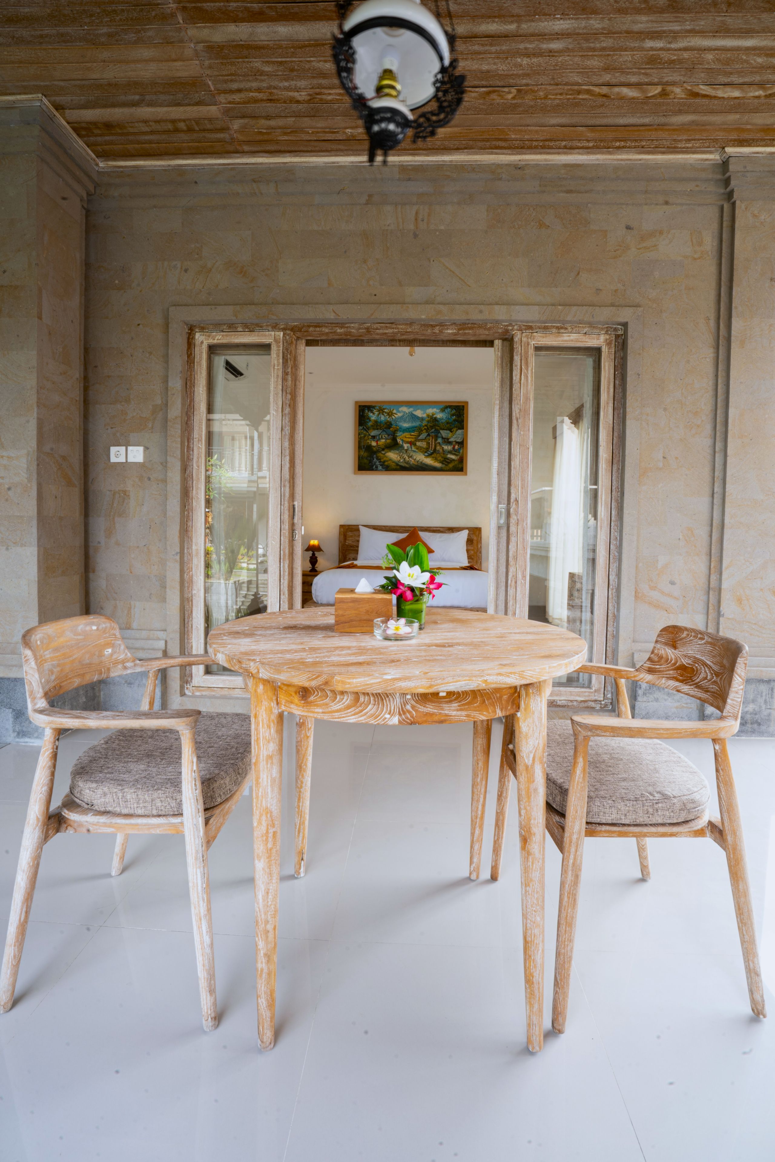 A room with a round wooden table, two chairs, a bed with a painting above it, and a ceiling light fixture.
