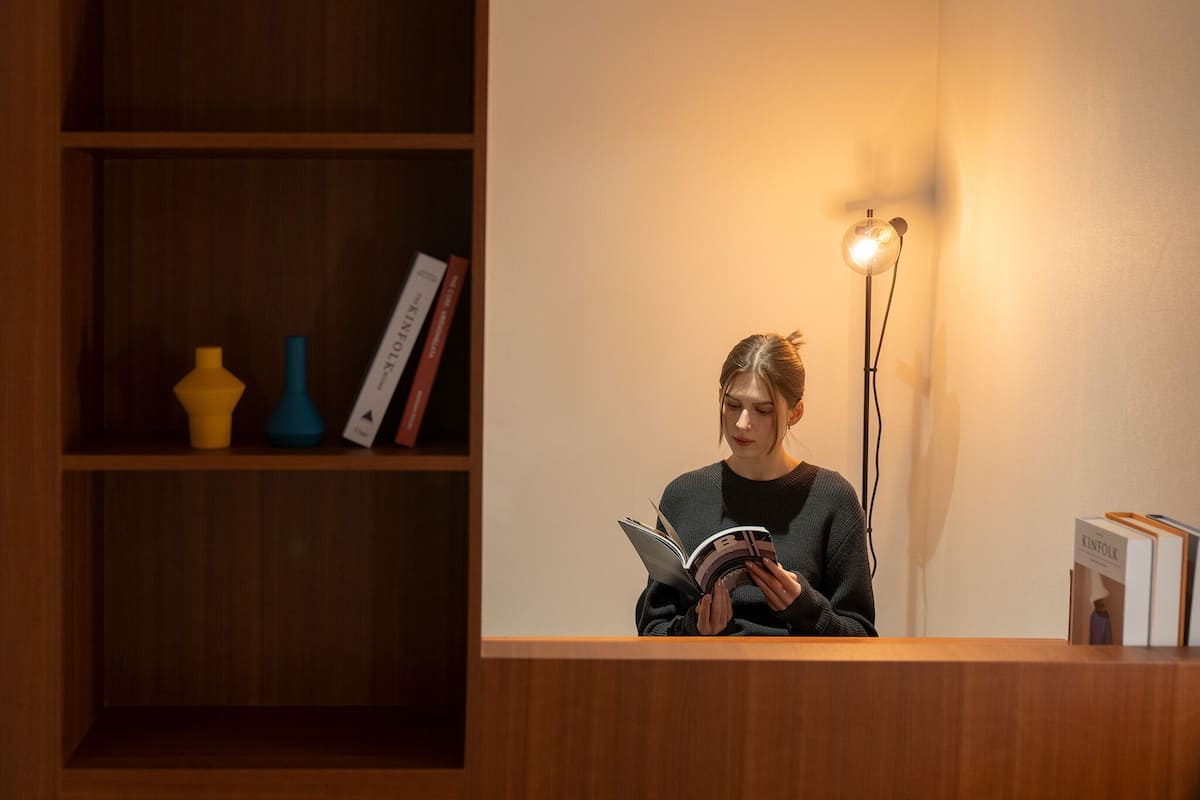 En person läser en bok i ett rum med en receptionsdisk i trä, en bokhylla med böcker och dekorativa föremål samt en golvlampa.