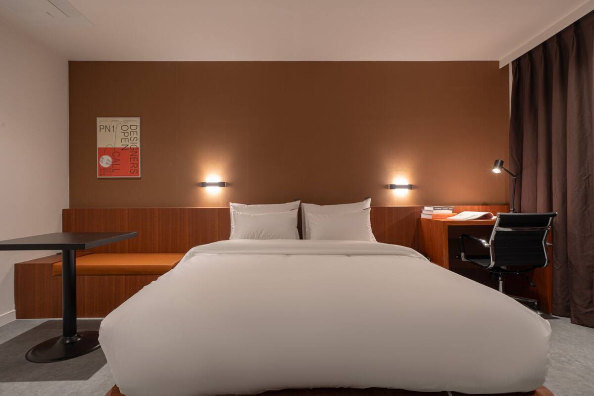 Ett hotellrum med en stor säng, ett skrivbord, en stol och två väggmonterade lampor.