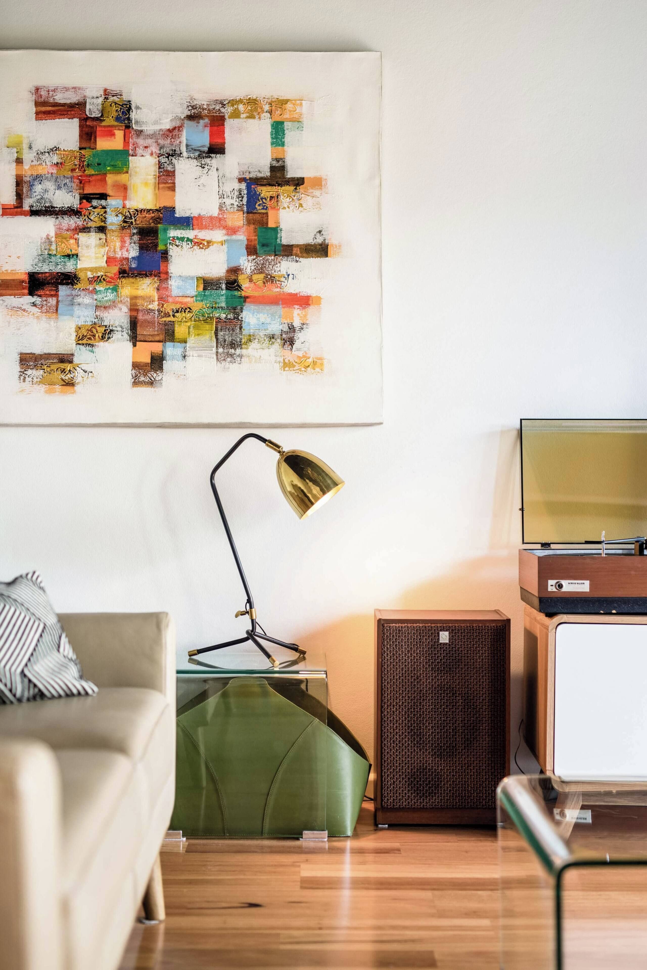 A living room with a beige sofa, a green ottoman, a floor lamp, a vintage television, and a wall-mounted abstract art piece.