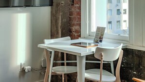 A room with a TV mounted on the wall, a table with chairs, a window with a view of buildings, and a brick wall.