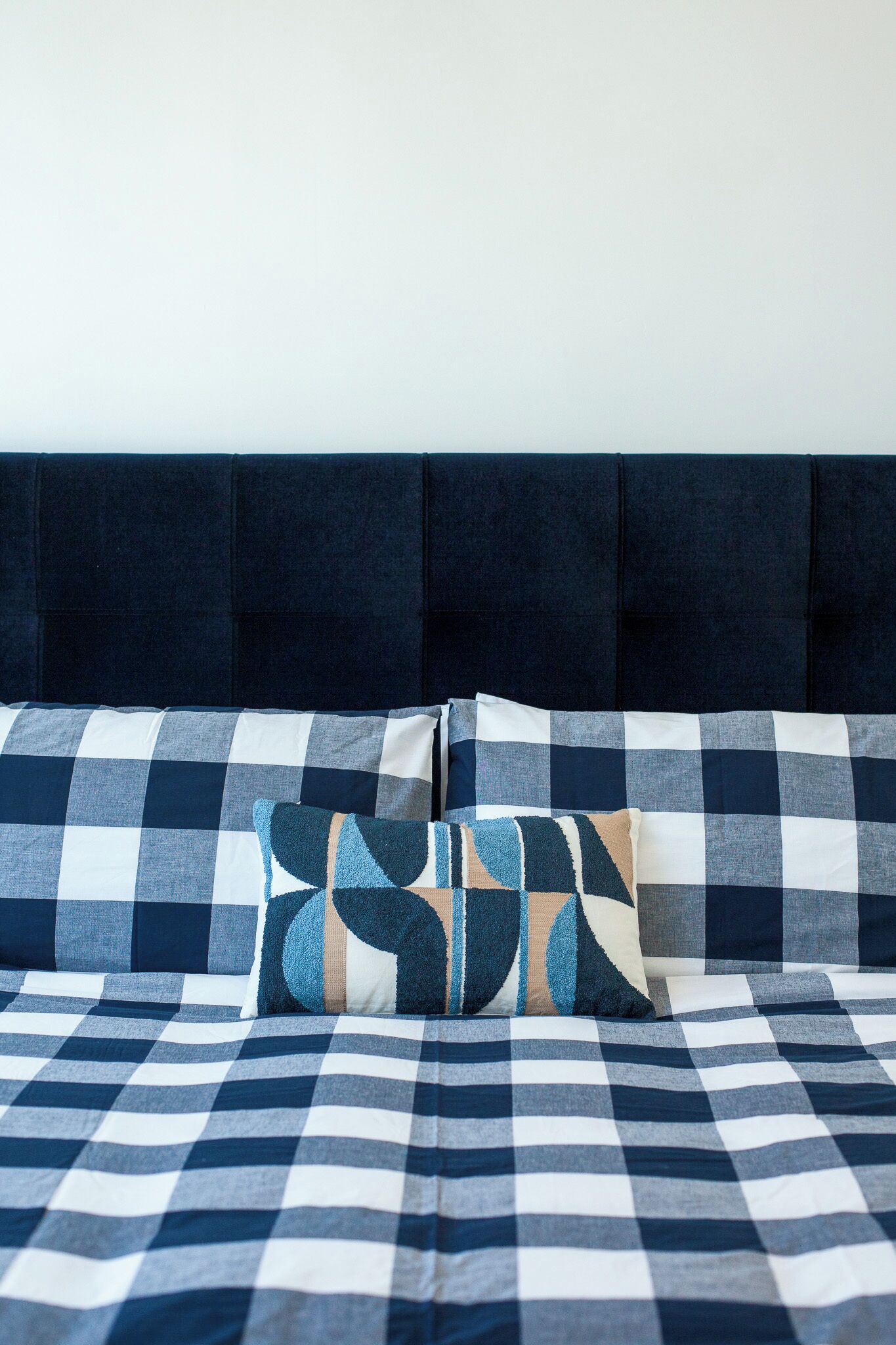 A bed with a checkered bedspread and two pillows, a dark headboard, and a plain wall.