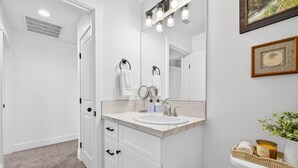 Ein Badezimmer mit einem weißen Waschbecken, Spiegel und Toilette. Handtücher hängen an der Wand, ein gerahmtes Bild und eine Pflanze stehen daneben.