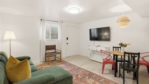 Una sala de estar con un sofá verde, una silla de madera, una mesa de comedor con sillas, un televisor en la pared y una alfombra estampada.