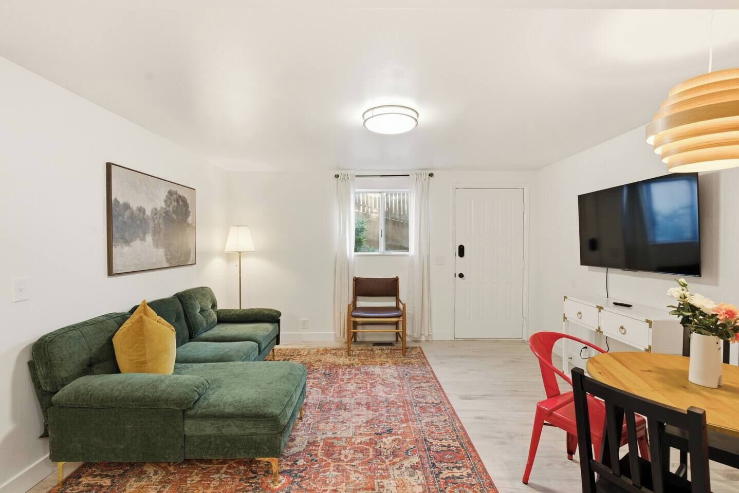 Una sala de estar con un sofá verde, un sillón rojo, una mesa de comedor de madera, un televisor montado en la pared y un tapete estampado.