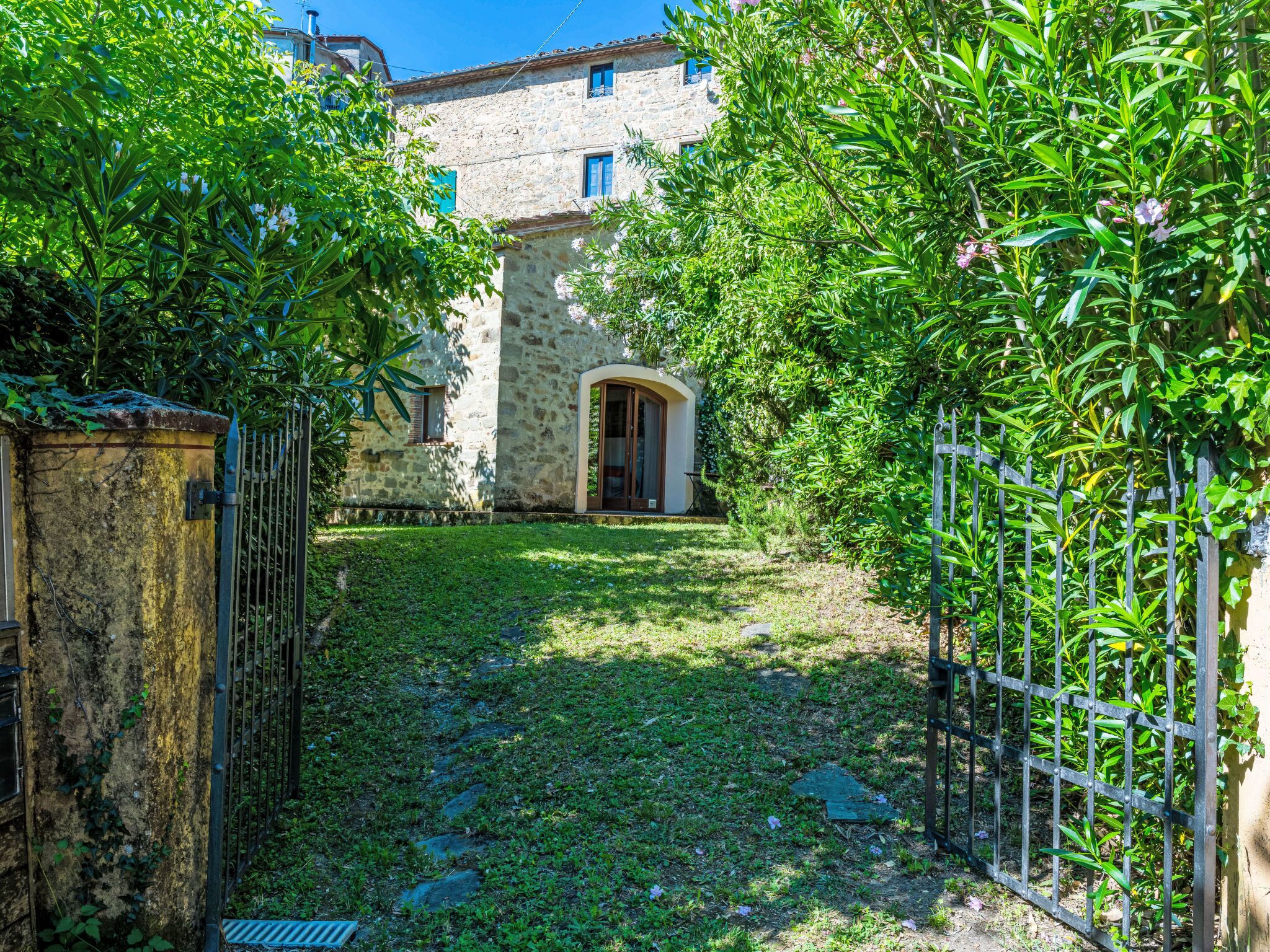 Un bâtiment en pierre avec une entrée donnant sur un jardin, un portail en métal et un chemin pavé menant à la porte.