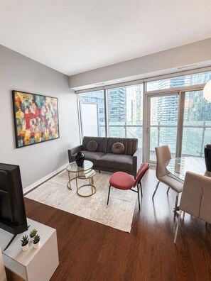 A modern living room with a sofa, coffee table, and chairs, a TV, and a large window with a city view. - 12 York | Modern 1BR Near CN Tower (Toronto)