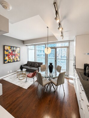 A modern apartment with a dining area, a gray sofa, a large window, and a kitchen. - 12 York | Modern 1BR Near CN Tower (Toronto)