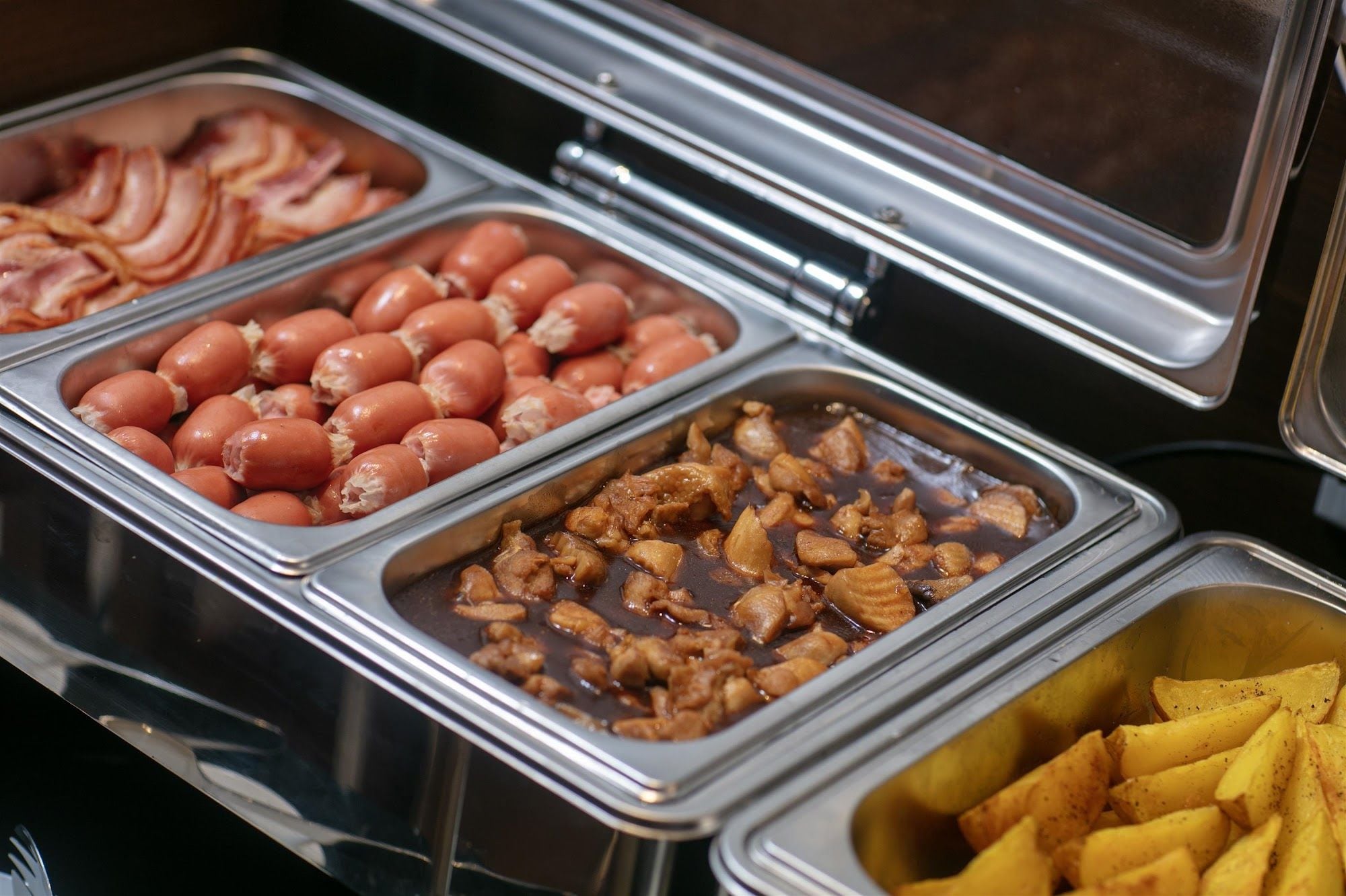 Un bufé con diversas opciones de comida, incluyendo salchichas, un plato de carne y papas fritas.