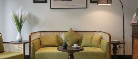 Una habitación con un sofá verde, una mesa redonda de madera, un florero con flores blancas y una lámpara.
