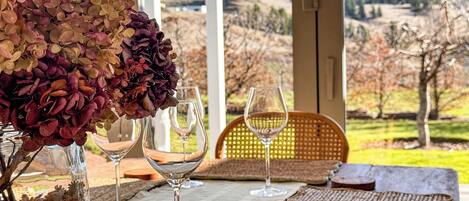 A table with a vase of dried flowers, two wine glasses, and a wicker chair with a view of a mountainous landscape.
