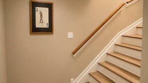 A staircase with wooden steps and a handrail, a framed playing card on the wall, and a light switch. - McCormick Meetup - Prime Location - Sleeps 16 (Chicago)
