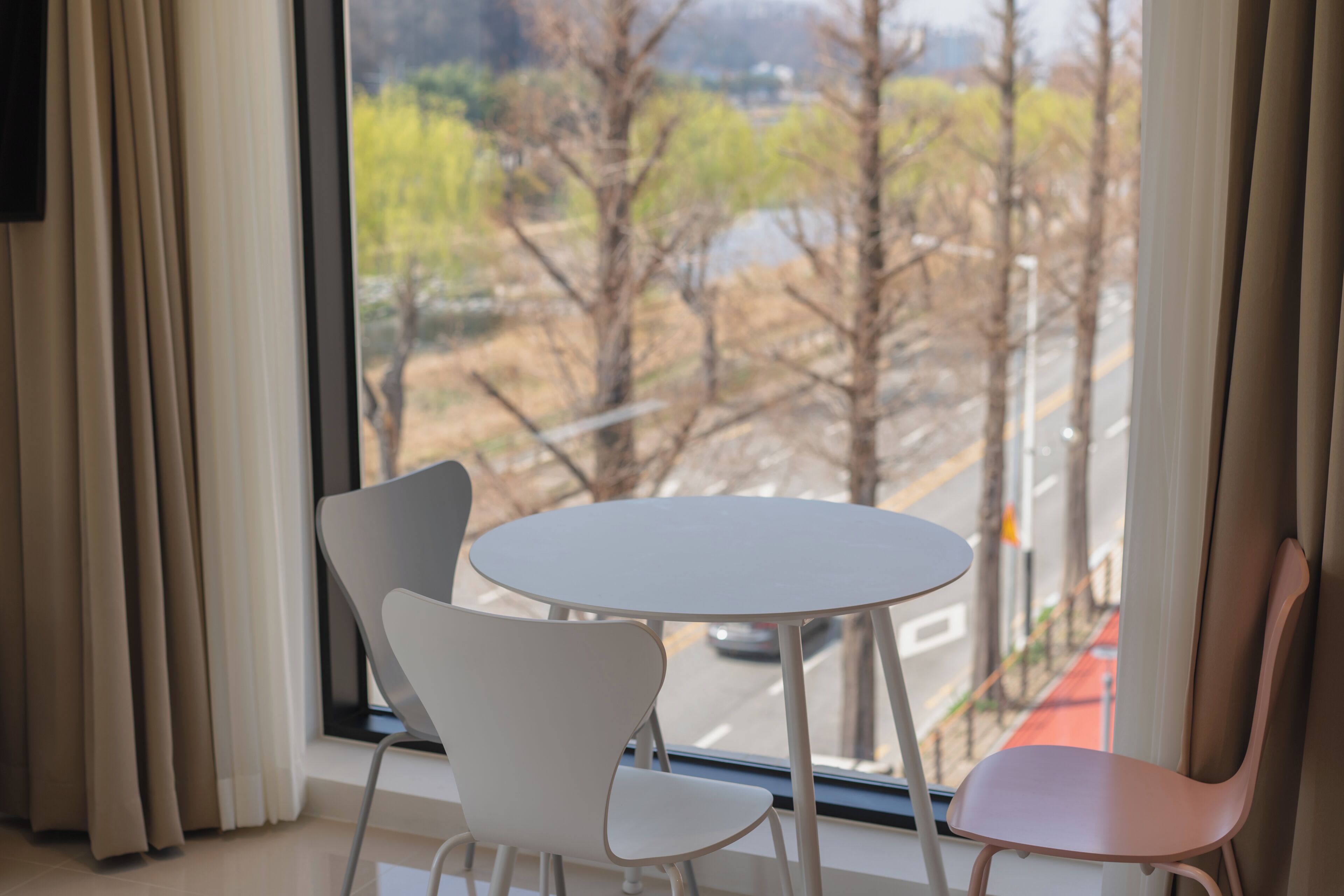 A room with a round table and two chairs, a window with curtains, and a view of a street and trees.