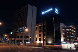 Un hotel de varios pisos con un llamativo letrero 'MH HOTEL', un edificio moderno con fachada de vidrio y un área de estacionamiento con bolardos.