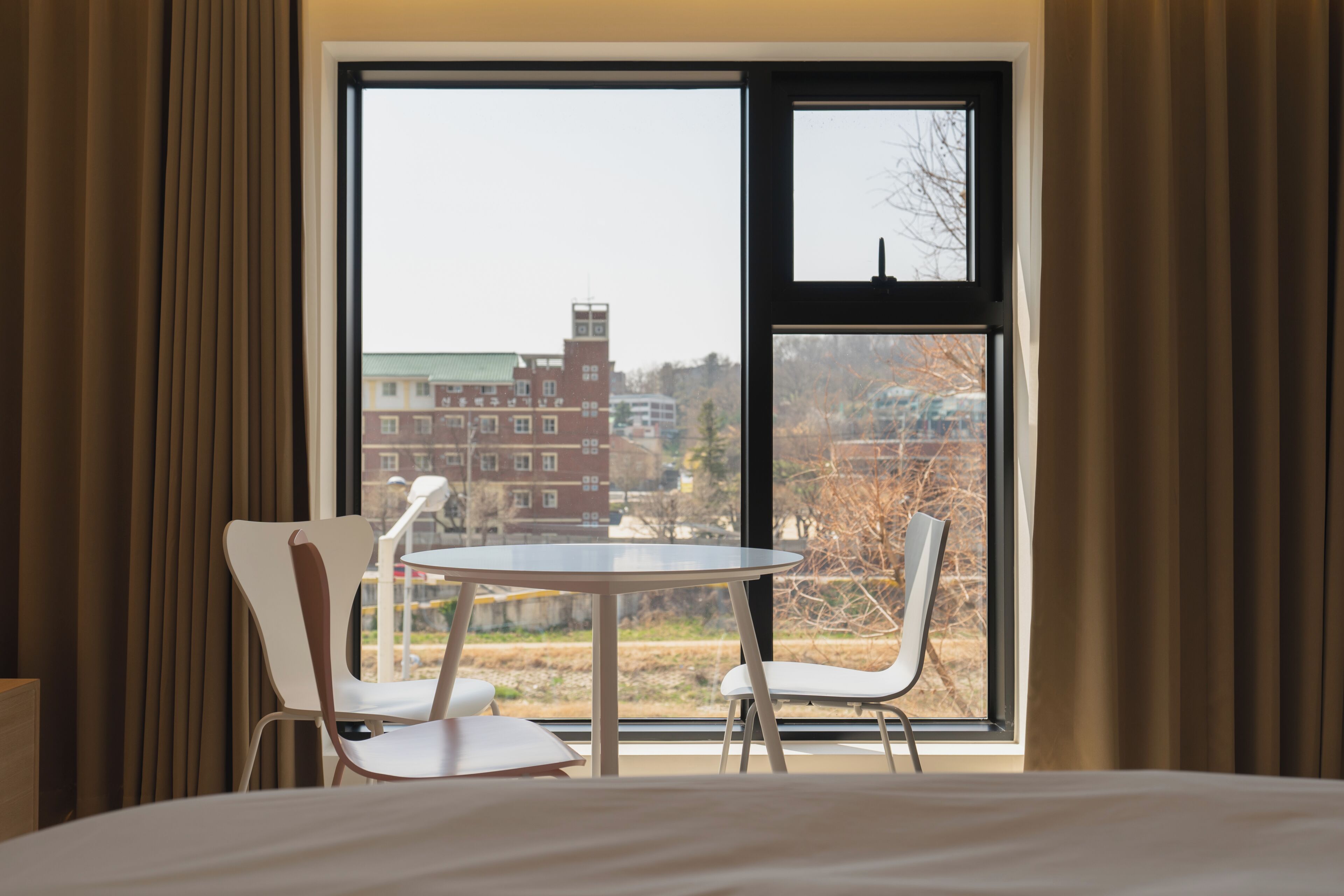 Habitación de hotel con un ventanal grande, dos sillas blancas, una mesita redonda y una cama.