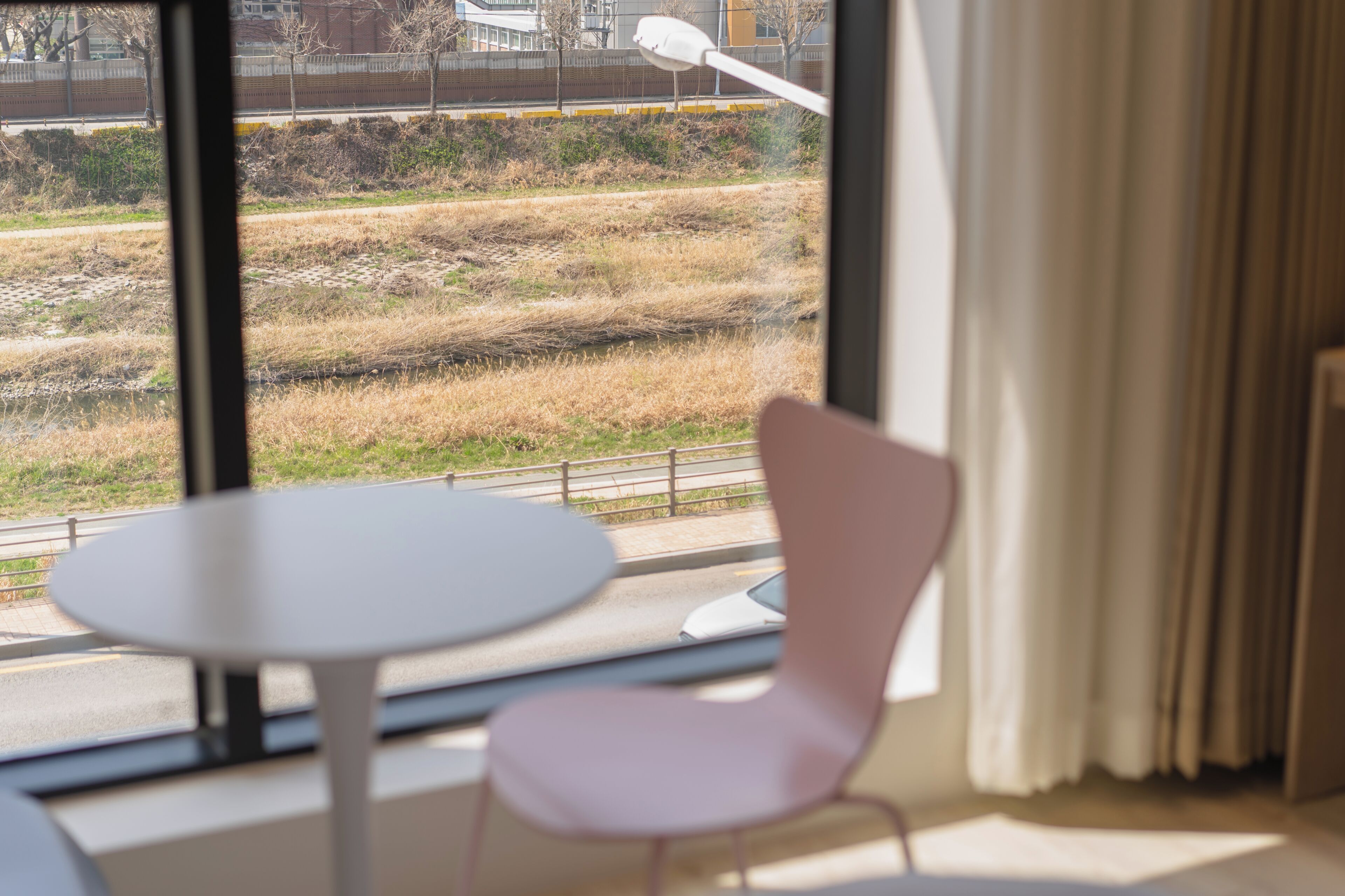 A room with a table and a chair, a window with a view of a grassy area and a fence, and a curtain on the window.