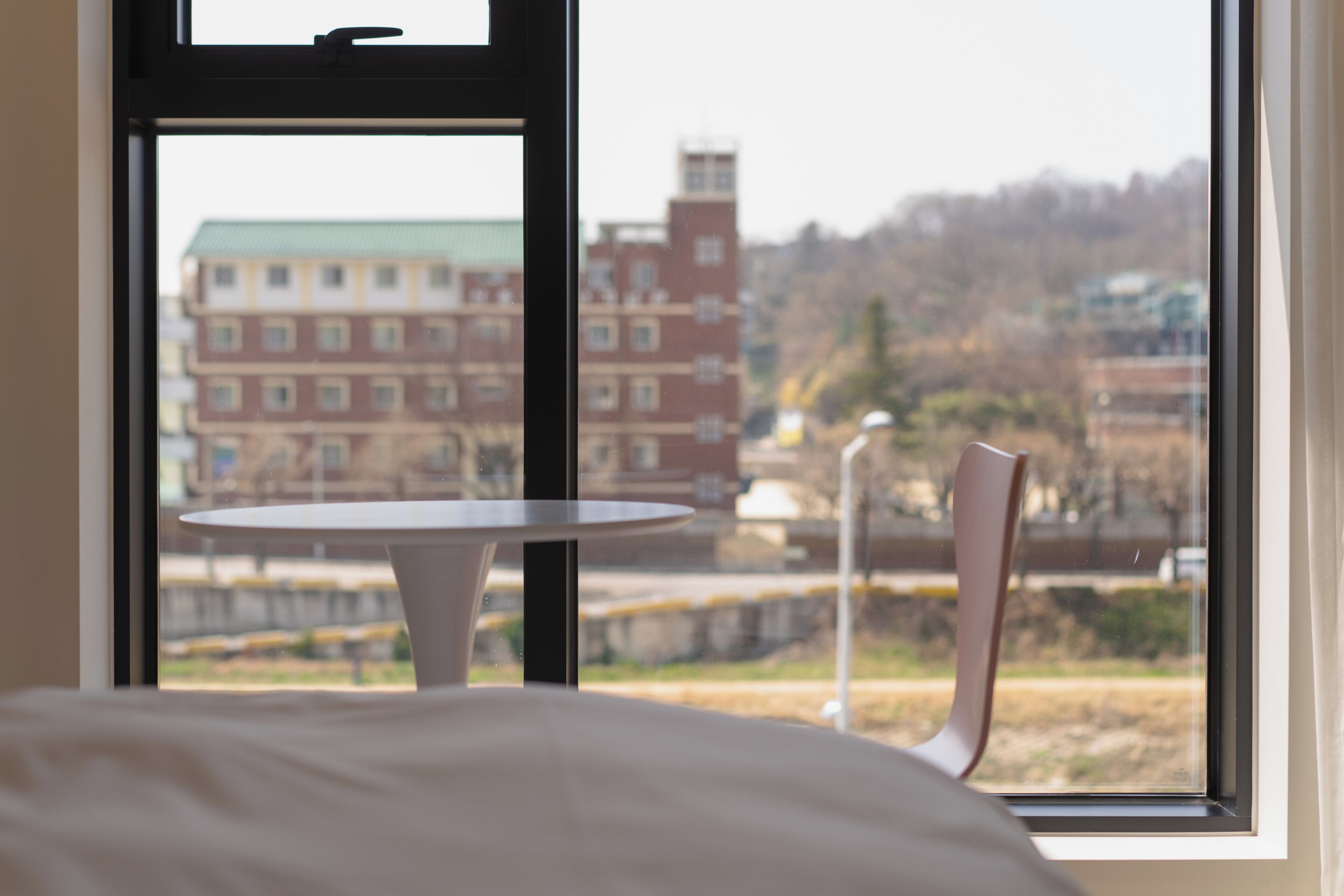 A room with a bed, a small round table, and a chair, with a window offering a view of buildings and a park.