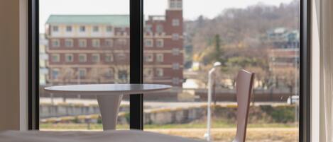 A room with a bed, a small round table, and a chair, with a window offering a view of buildings and a park.