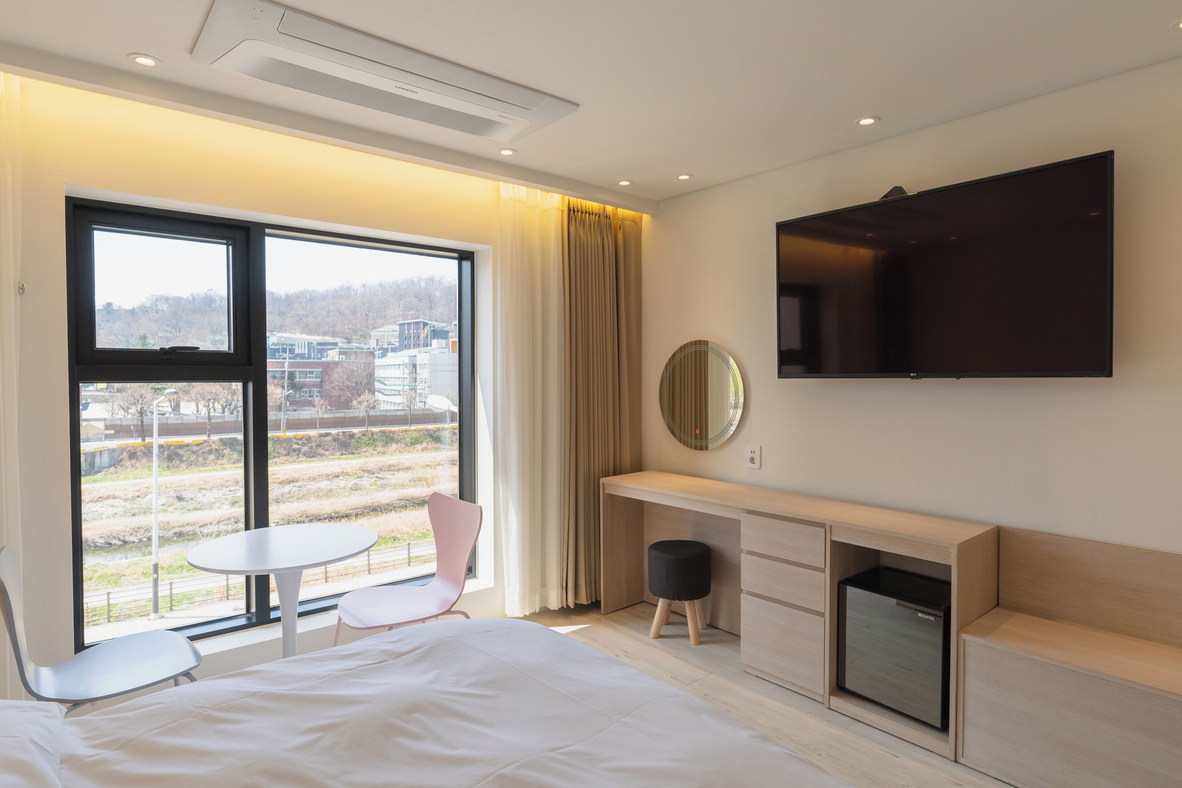 A modern hotel room with a bed, a desk, a chair, a small table, a TV, and a view of the outside.