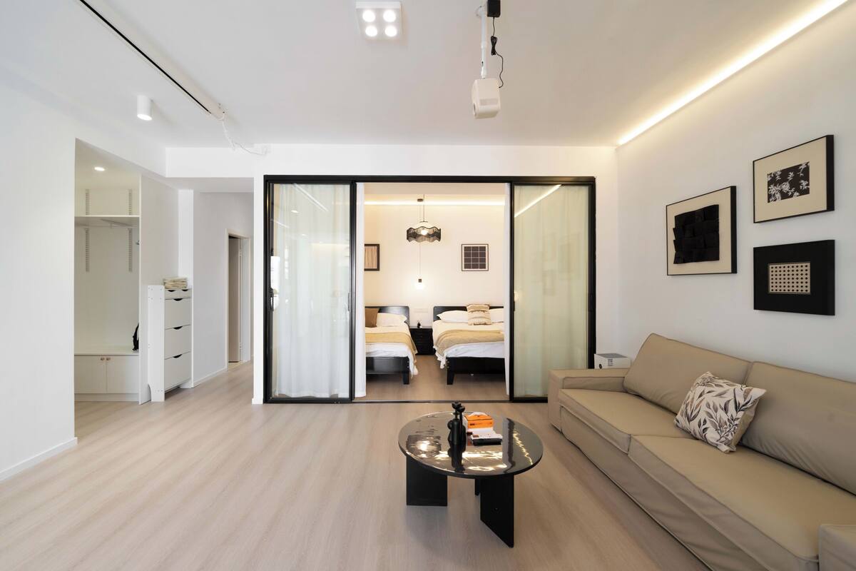 A modern hotel room with a glass partition, a sofa, a coffee table, and a bed with a nightstand.