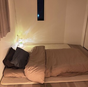Ein Einzelbett mit beiger Matratze und Kissen, ein kleines Fenster und eine Wandleuchte.