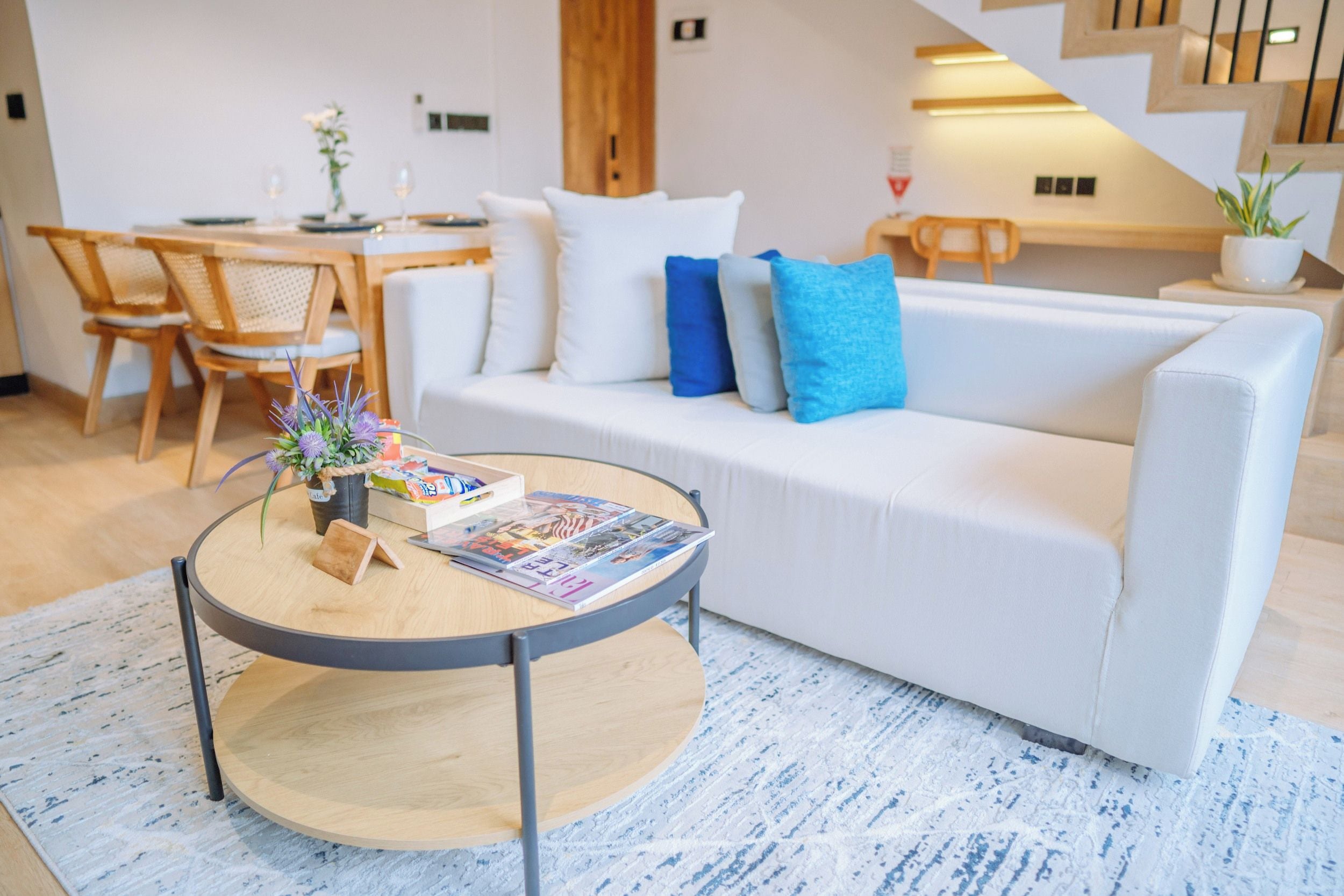 A modern living room with a white sofa, a round coffee table with magazines, and a dining area with wooden furniture.