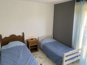 Ein Schlafzimmer mit zwei Einzelbetten, einem Nachttisch mit Lampe und einem Fenster mit durchsichtigen Vorhängen.