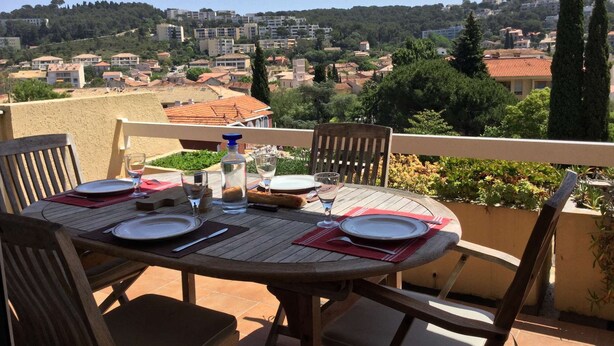 A round wooden table set for two with plates, cutlery, and glasses, overlooking a hillside town. - Vacation rental 2 bedrooms, air conditioning, Wi-Fi, balcony, barbecue, private parking (ST MANDRIER SUR MER)