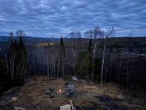 A forested area with a fire pit and chairs, surrounded by trees and a distant view of a town.
