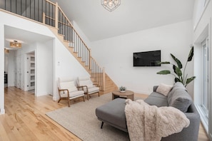 A modern living room with a staircase, a grey sofa, a white armchair, and a TV mounted on the wall.