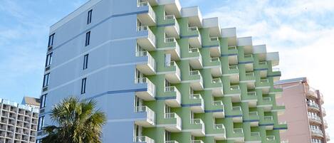 A multi-story hotel with a distinctive green and white facade, balconies, and a palm tree in the foreground.