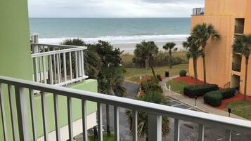 A balcony with a view of the beach and ocean, a parking area, and a building with a green and yellow facade.
