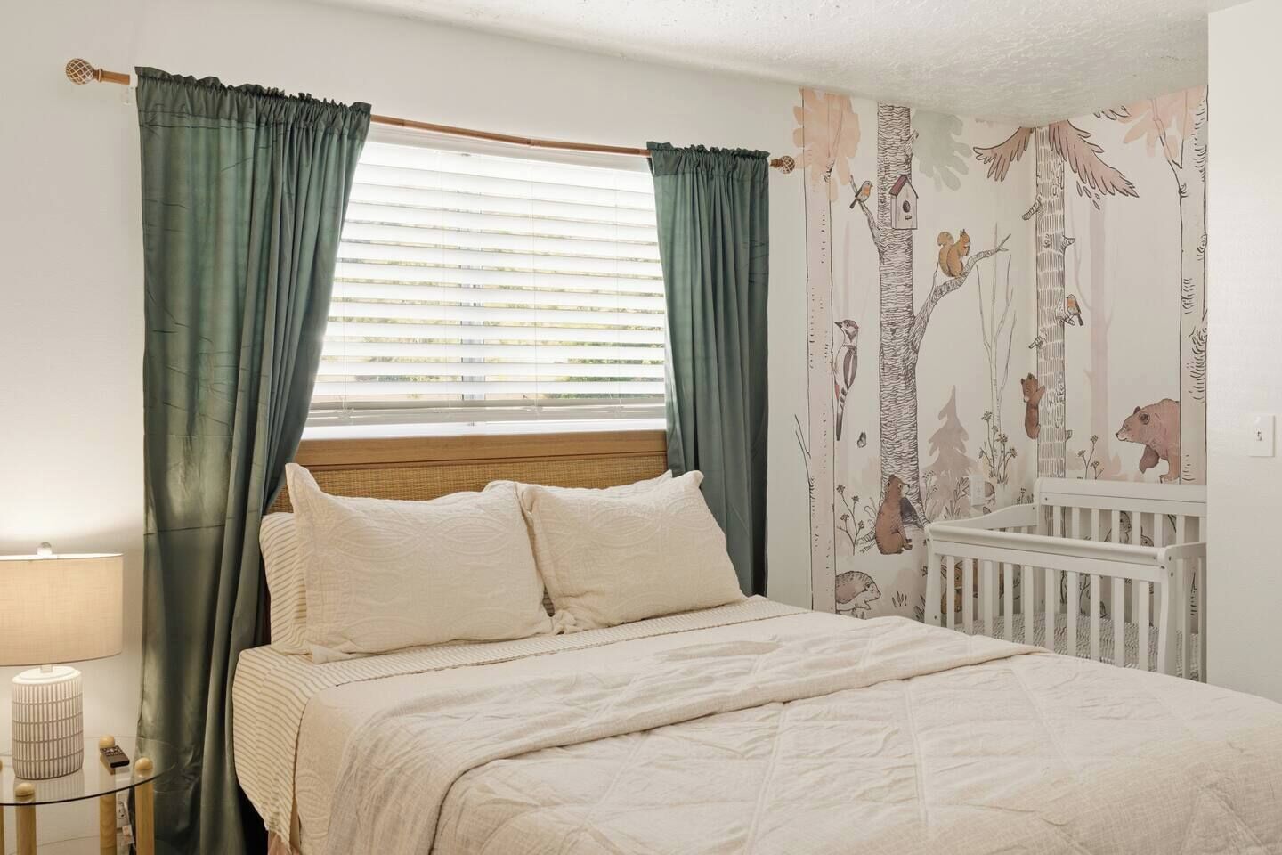 Une chambre à coucher comprenant un lit, des rideaux, une table de chevet, une lampe et un mur orné d’une fresque sur le thème de la nature.