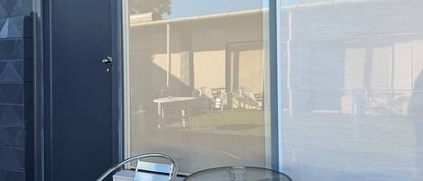 Un patio con dos sillas de metal y una mesa de vidrio, una puerta oscura y vistas a un espacio exterior con mesas y sillas.