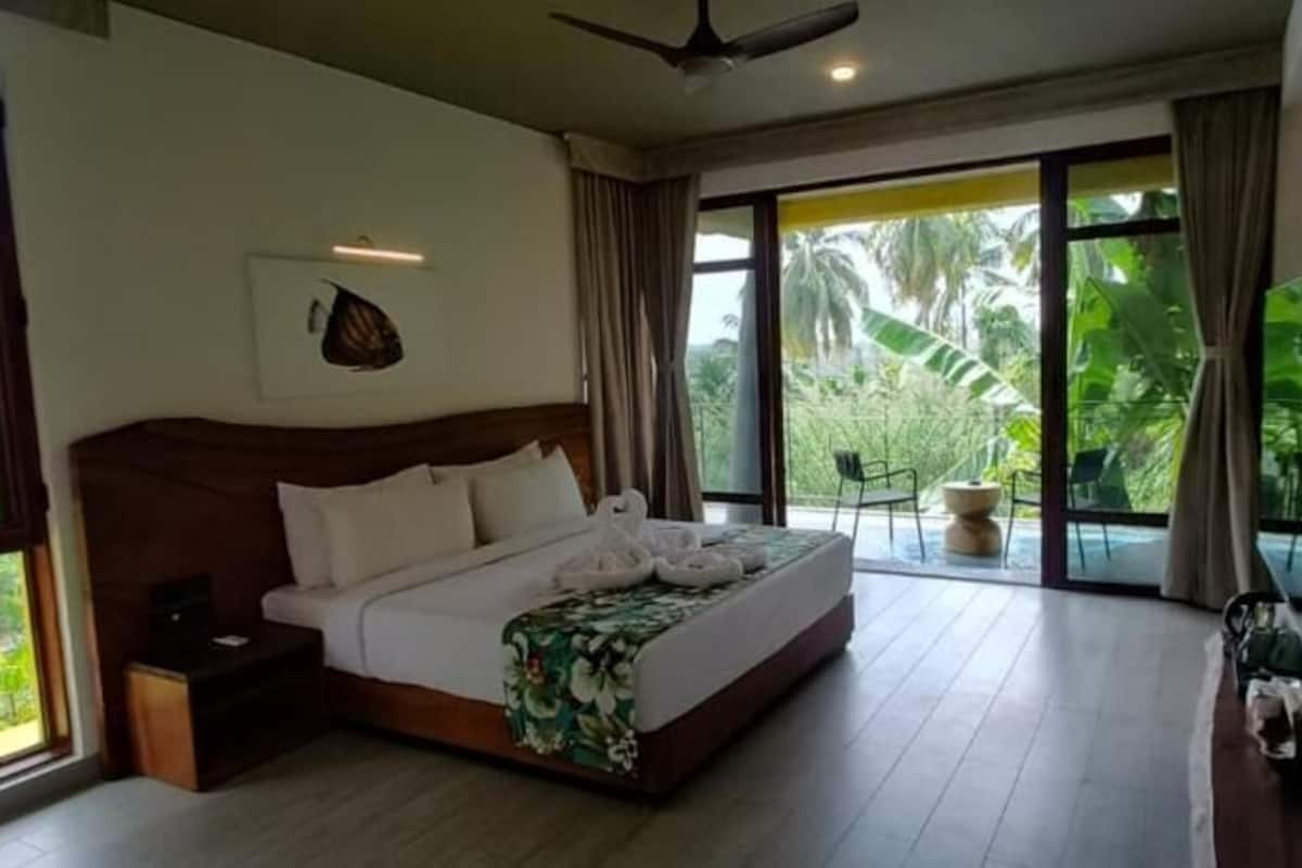 A spacious bedroom with a large bed, a ceiling fan, a TV, and a balcony with a view of greenery.
