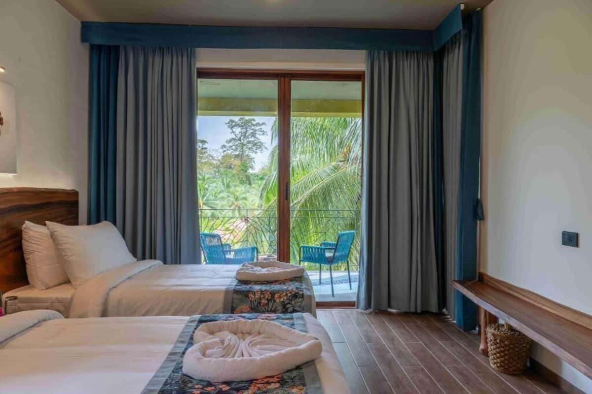 A hotel room with two beds, a balcony with chairs, and a view of greenery.