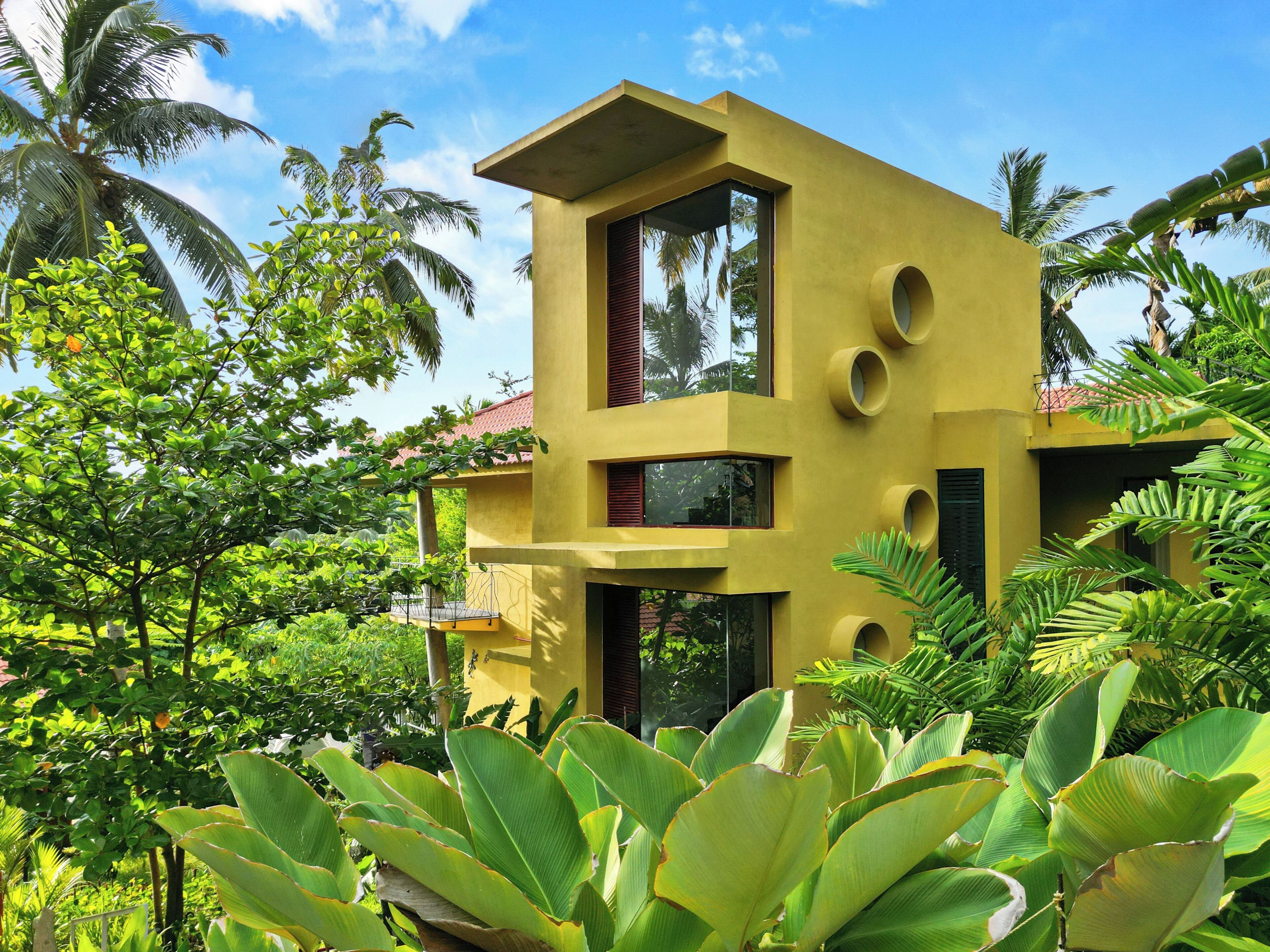 A modern yellow building with a balcony, surrounded by lush greenery and palm trees.