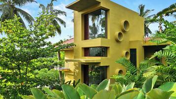 A modern yellow building with a balcony, surrounded by lush greenery and palm trees.