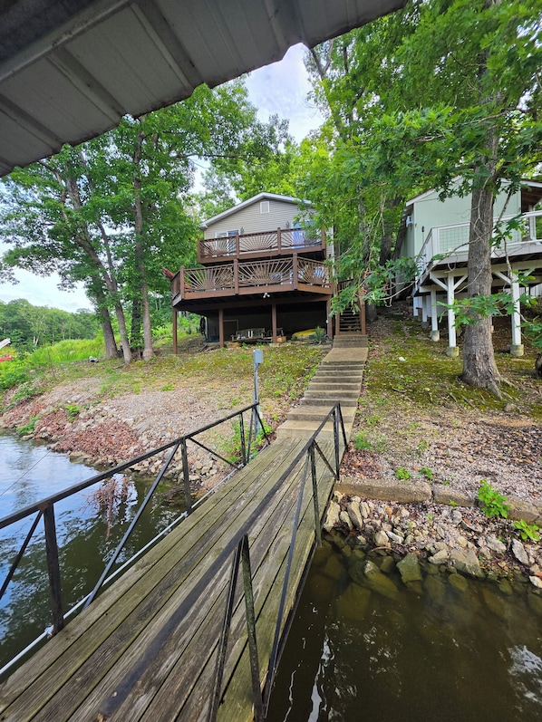 A wooden dock leading to a house with a balcony, surrounded by trees and a body of water. - Lakefront Cove Retreat, Private Dock, Huge Deck (Rocky Mount)